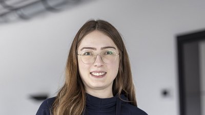 Eine junge Frau mit langen, braunen Haaren und Brille lächelt freundlich in einem modernen, hellen Raum.