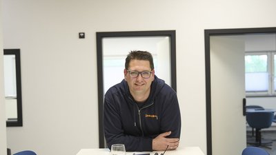 Ein Mann mit Brille lächelt freundlich, während er an einem Tisch in einem modernen Büro steht, umgeben von neutralen Farben.