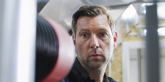 Ein Mann mit ernstem Gesichtsausdruck steht in einem modernen Fitnessstudio, umgeben von Trainingsgeräten und Gewichten.