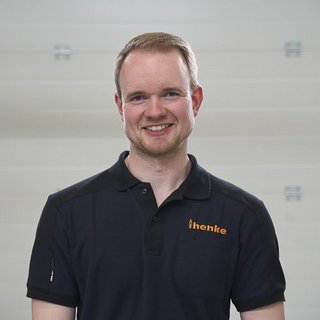 Ein lächelnder Mann in einem schwarzen Poloshirt mit dem Logo 'henke' steht vor einer hellen, neutralen Wand.