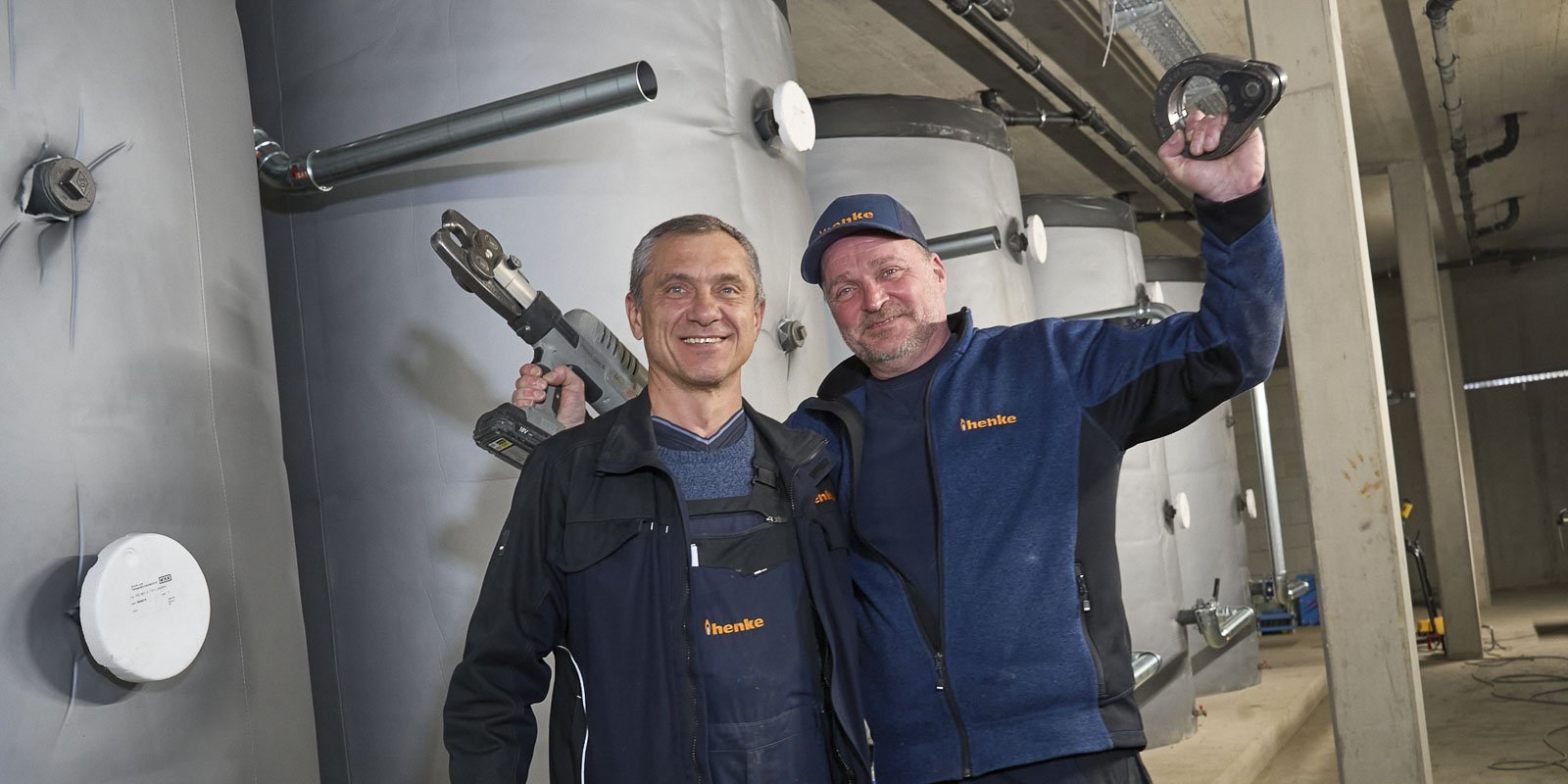 Zwei Männer stehen in einem industriellen Raum, lächeln und halten Werkzeuge in der Hand, umgeben von großen silbernen Tanks.