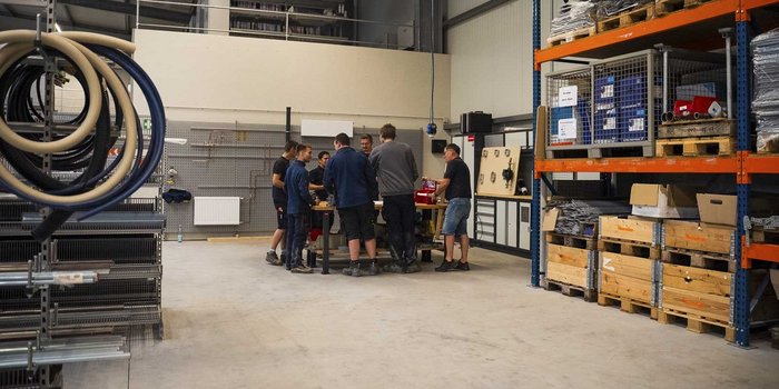 In einer hellen Werkstatt arbeiten mehrere Männer zusammen, um an einem Projekt zu tüfteln, umgeben von Regalen voller Materialien.