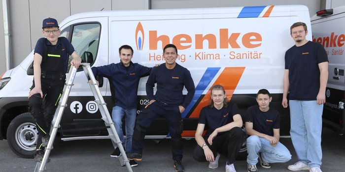 Die Gruppe von Handwerkern zeigt sich vor einem Fahrzeug mit dem Logo 'henke', während sie eine Leiter benutzen.
