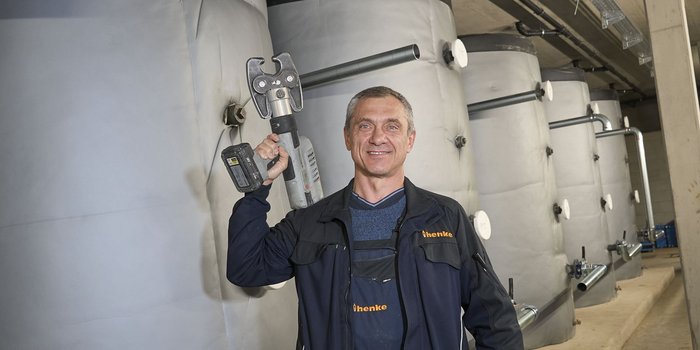 Ein lächelnder Handwerker hält ein Werkzeug in einer Industrieumgebung mit großen, grauen Tanks im Hintergrund.