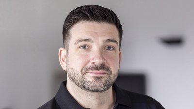 Der Mann trägt ein schwarzes Polohemd und hat einen neutralen Gesichtsausdruck, umgeben von einem hellen Hintergrund.