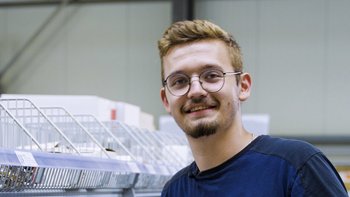 Ein junger Mann mit Brille lächelt freundlich, während er in einem modernen Lager mit Regalen steht.