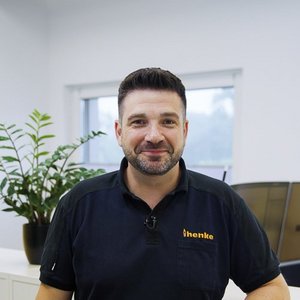 Ein Mann mit dunklem Haar und Bart lächelt freundlich in einem modernen Büro mit Pflanzen im Hintergrund.
