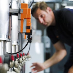 Im Hintergrund sind glänzende Metallrohre zu sehen, während ein Mann mit einem orangefarbenen Gerät an der Steuerung arbeitet.