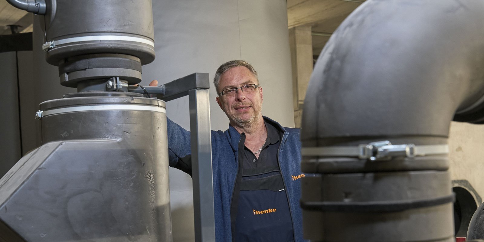 In einer Fabrikumgebung posiert ein Mann mit Brille, der an einer großen Rohrleitung arbeitet und freundlich in die Kamera schaut.