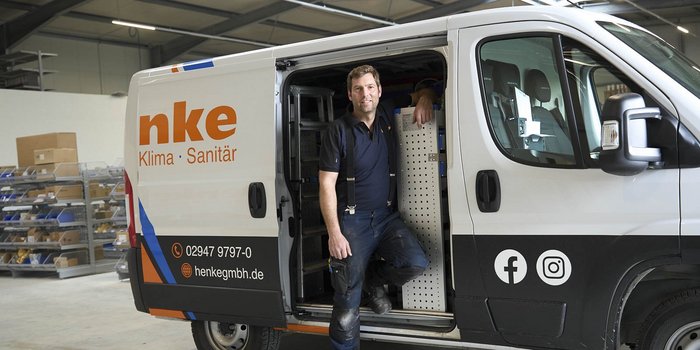 Ein Handwerker steht lächelnd in einem Lieferwagen, umgeben von Werkzeugen und Materialien für Klima- und Sanitärarbeiten.