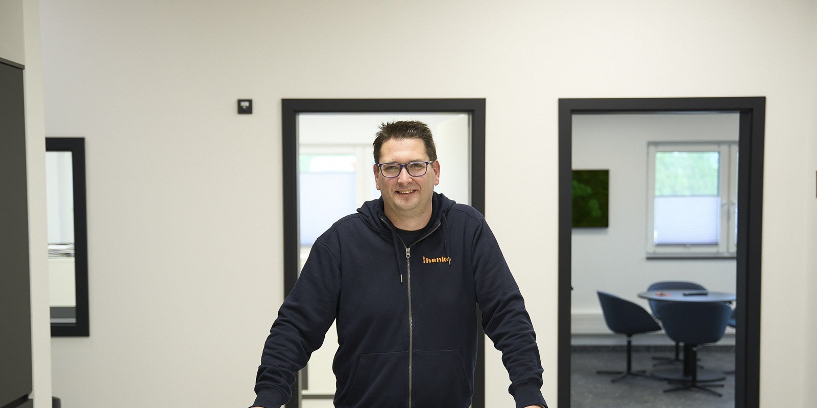 Ein Mann mit Brille und Kapuzenpullover lächelt freundlich, während er an einem Tisch in einem modernen Büro steht.