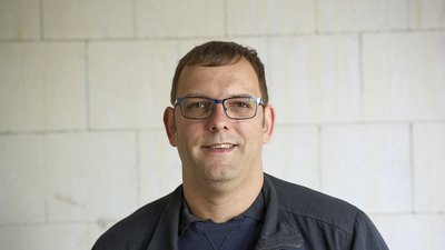 Ein Mann mit Brille lächelt freundlich vor einer hellen, strukturierten Wand aus Mauersteinen.