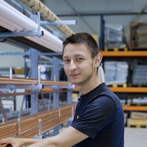 Ein junger Mann mit kurzem Haar lächelt, während er an einer Produktionslinie mit Kupferrohren arbeitet.