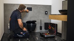 Ein Handwerker kniet in einem modernen Badezimmer und installiert eine schwarze Toilettenspülung an der Wand.