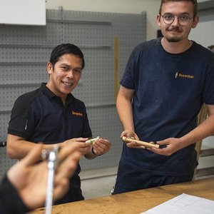 In einer hellen Werkstatt zeigen zwei Männer stolz ihre handgefertigten Holzobjekte, während sie freundlich in die Kamera blicken.