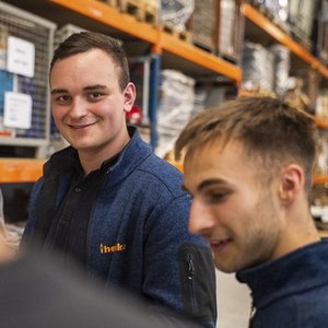 Zwei Männer in blauen Arbeitsjacken unterhalten sich in einem Lager, während ein Kollege lächelt und in die Kamera schaut.