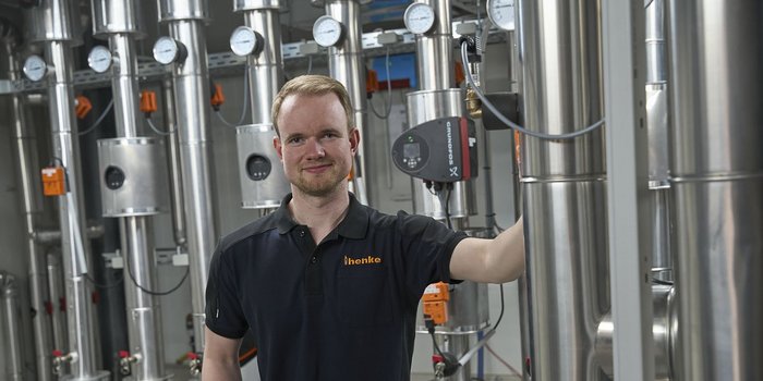 Ein Mann in einem schwarzen Hemd steht neben glänzenden Maschinen mit Druckmessgeräten und lächelt freundlich in die Kamera.