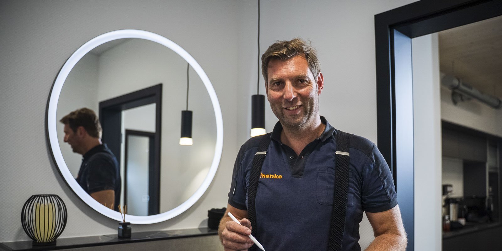 Ein lächelnder Mann mit einem Stift in der Hand steht in einem modernen Raum mit einem großen runden Spiegel.