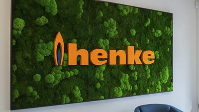 Eine moderne Wand mit lebendigem Moos und dem leuchtenden Schriftzug 'henke' in Orange, umgeben von stilvollen blauen Stühlen.