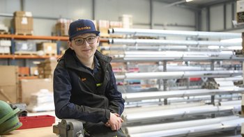 Ein junger Mann sitzt in einem Lager, umgeben von Regalen voller Materialien und Werkzeugen, mit einem freundlichen Lächeln.