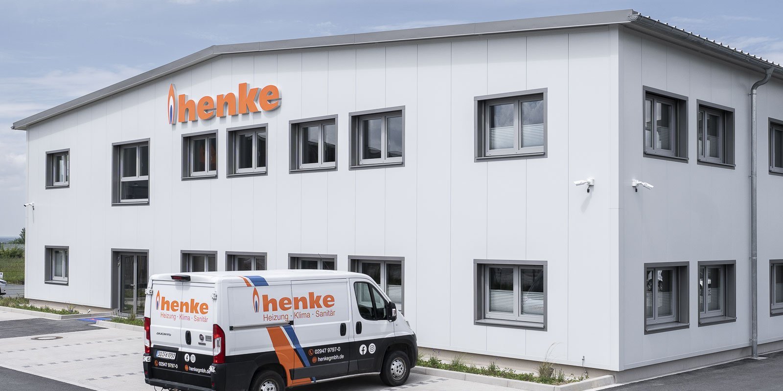 Das Gebäude von 'henke' zeigt große Fenster und eine helle Fassade, während ein Firmenfahrzeug davor parkt.