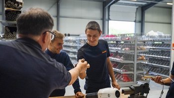 Fünf Männer in Arbeitskleidung stehen an einem Tisch und tauschen Werkzeuge aus, während sie lächeln und sich unterhalten.