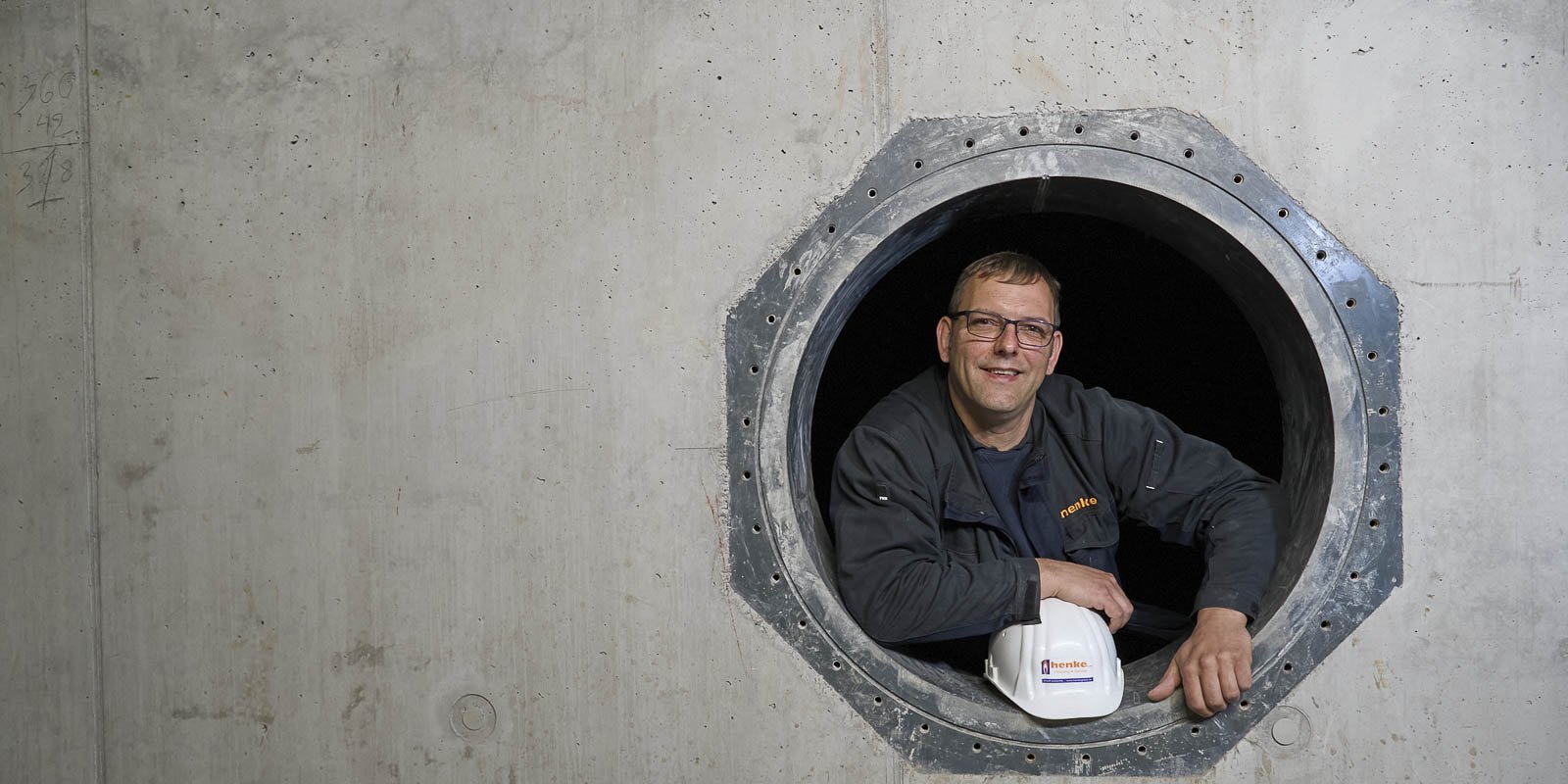 Ein Bauarbeiter lächelt fröhlich aus einem großen runden Fenster in einer Betonwand, während er einen Schutzhelm hält.