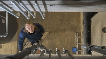 Ein junger Mann schaut von einer Leiter aus nach oben, umgeben von Rohren und einer grauen Wand in einem industriellen Raum.