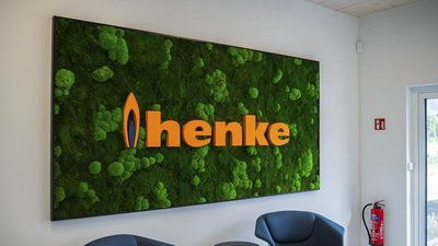 Ein modernes Wandbild mit dem Schriftzug 'henke' in leuchtendem Orange, umgeben von lebendigem, grünem Moos.