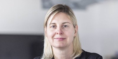 Eine Frau mit blonden Haaren lächelt sanft in die Kamera, während sie in einem modernen Raum steht.