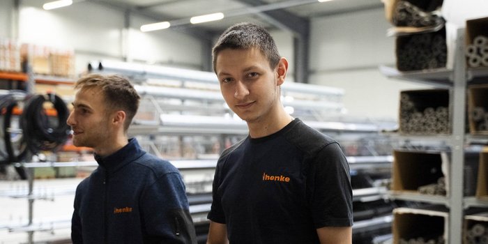 Zwei junge Männer stehen in einer modernen Werkstatt, umgeben von Metallrohren und Werkzeugen, mit freundlichen Gesichtsausdrücken.